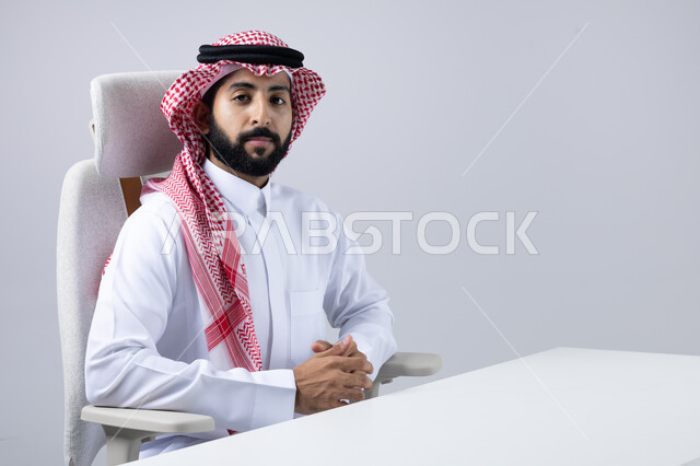 مفهوم الرجولة والقوة والثقة بالنفس، النظر إلى الكاميرا بايماءات الجدية والسرور، الاهتمام بالأناقة والمظهر الخارجي، بورتريه مقرب لرجل عربي خليجي سعودي يرتدي الثوب التقليدي والشماغ يجلس بأيدي متشابكة بتعابير الحزم، خلفية بيضاء