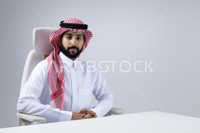 The concept of masculinity, strength and self-confidence, looking at the camera with gestures of seriousness and pleasure, interest in elegance and outward appearance, close-up portrait of a Saudi Arabian Gulf man wearing a traditional thobe and shemagh sitting with clasped hands with an expression of firmness, white background