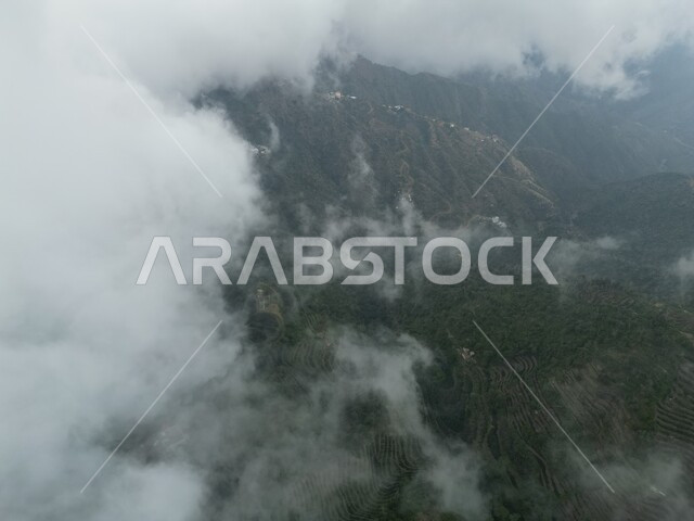 Cloudy sky with white clouds over the mountains of Fifa, natural vegetation on the mountain peaks and heights, green agricultural terraces in Jazan in the Kingdom of Saudi Arabia, natural tourist places and landmarks in Saudi Arabia, nature background
