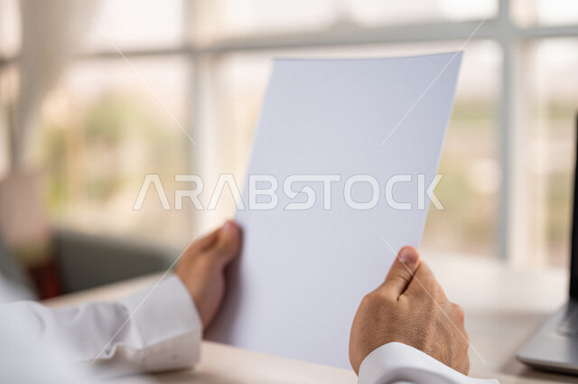 صورة مقربة ليد رجل عربي خليجي سعودي يحمل اوراق خاصة بالعمل، أوراق بيضاء فارغة، موظف يجلس أمام الحاسوب المحمول، سكرتير يدير الاعمال الورقية، أعمال مكتبية