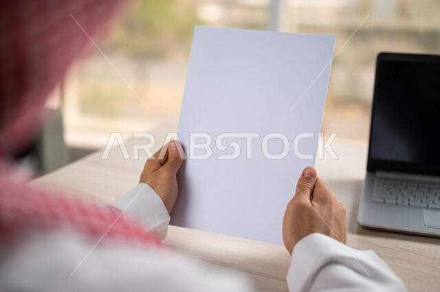 صورة مقربة ليد رجل عربي خليجي سعودي يحمل اوراق خاصة بالعمل، أوراق بيضاء فارغة، موظف يجلس أمام الحاسوب المحمول، سكرتير يدير الاعمال الورقية، أعمال مكتبية