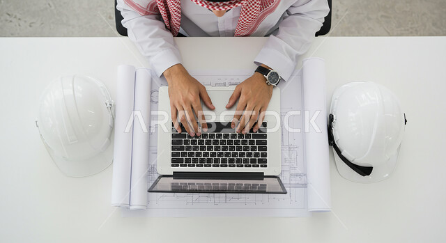  مفهوم ادارة المشاريع الهندسية والانشاءات، دمج التقنية والتكنولوجيا في مجال الهندسة، العمل في القطاع الهندسي في المملكة العربية السعودية، مهنة ووظيفة شبابية، صورة علوية لرجل عربي خليجي سعودي يرتدي الشماغ والثوب التقليدي يعمل على الحاسوب المحمول