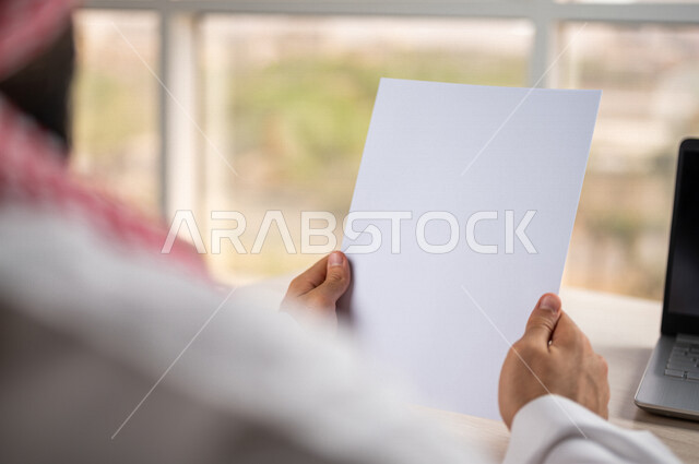 Close-up of the hand of a Saudi Arabian Gulf man holding work papers, blank white papers, an employee sitting in front of a laptop, a secretary managing paperwork, office work