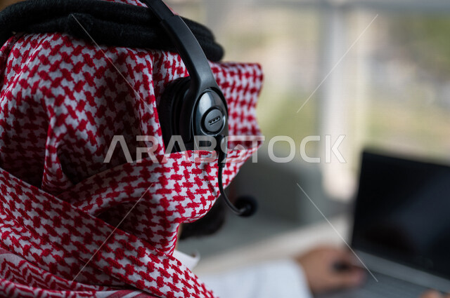 صورة مقربة ليد رجل عربي خليجي سعودي يعمل في خدمة العملاء، موظف يرتدي سماعة الاتصال، استعلامات واستفسارات، الاجابه على اسئلة العملاء، عقد اجتماع عمل عبر الانترنت، العمل عن بعد من خلال الحاسوب المحمول، متابعة وتطوير العمل