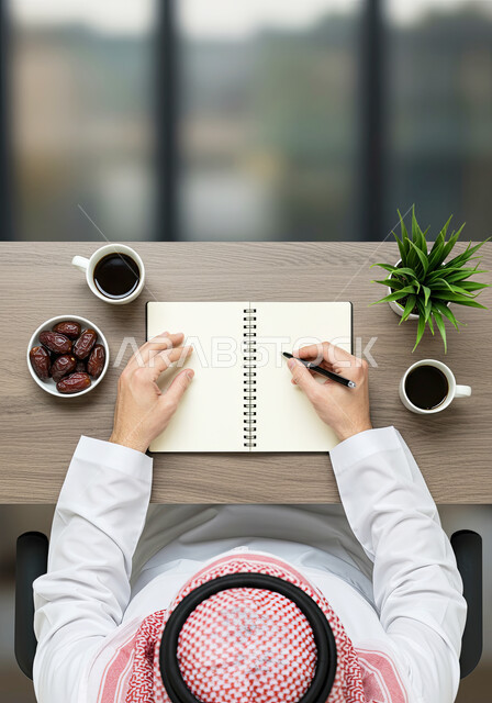 احتراف الكتابة بفنون الخط العربي، صورة علوية لرجل خطاط عربي خليجي سعودي يرتدي الثوب التقليدي والشماغ يقوم بالتخطيط باستخدام القلم، تعليم مهارات الرسم بالخطوط العربية، شرب القهوة العربية الساخنة، تناول حبات التمر الطازجة