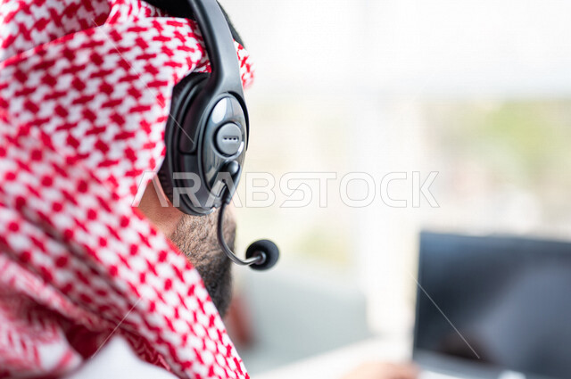 Close-up of the hand of a Saudi Arabian Gulf man working in customer service, an employee wearing a headset, inquiries and inquiries, answering customer questions, holding a business meeting via the Internet, working remotely through a laptop, following u