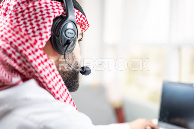 صورة مقربة ليد رجل عربي خليجي سعودي يعمل في خدمة العملاء، موظف يرتدي سماعة الاتصال، استعلامات واستفسارات، الاجابه على اسئلة العملاء، عقد اجتماع عمل عبر الانترنت، العمل عن بعد من خلال الحاسوب المحمول، متابعة وتطوير العمل