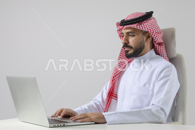 مفهوم دمج العمل مع التقنيه والتكنولوجيا, إنجاز مهام العمل عبر جهاز الابتوب, بورتريه لرجل عربي خليجي سعودي مبتسم يرتدي الثوب والشماغ التقليدي يعمل على الحاسب المحمول  بإيماءات السرور, خلفيه بيضاء