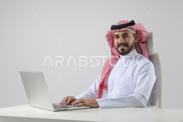 مفهوم دمج العمل مع التقنيه والتكنولوجيا, إنجاز مهام العمل عبر جهاز الابتوب, بورتريه لرجل عربي خليجي سعودي مبتسم يرتدي الثوب والشماغ التقليدي يعمل على الحاسب المحمول  بإيماءات السرور, خلفيه بيضاء