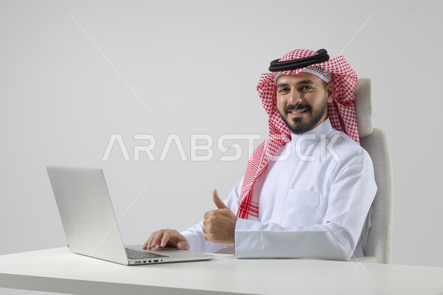 تعابير الاعجاب والقبول، مفهوم دمج العمل مع التقنيه والتكنولوجيا, إنجاز مهام العمل عبر جهاز الابتوب, بورتريه لرجل عربي خليجي سعودي مبتسم يرتدي الثوب والشماغ التقليدي يعمل على الحاسب المحمو بإيماءات السرور, خلفيه بيضاء