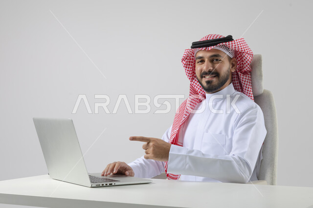 مفهوم دمج العمل مع التقنيه والتكنولوجيا, إنجاز مهام العمل عبر جهاز الابتوب, بورتريه لرجل عربي خليجي سعودي مبتسم يرتدي الثوب والشماغ التقليدي يعمل على الحاسب المحمول  بإيماءات السرور, خلفيه بيضاء
