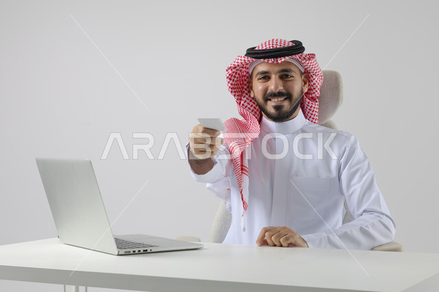 عرض موك اب ابيض صغير، مفهوم دمج العمل مع التقنيه والتكنولوجيا, إنجاز مهام العمل عبر جهاز الابتوب, بورتريه لرجل عربي خليجي سعودي مبتسم يرتدي الثوب والشماغ التقليدي يمسك بيده بطاقة تعريفية، العمل على الحاسب المحمول  بإيماءات السرور, خلفيه بيضاء