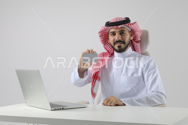 Small white mockup, concept of integrating work with technology, completing work tasks via laptop, portrait of a smiling Saudi Arabian Gulf man wearing traditional thobe and shemagh holding an ID card, working on laptop with gestures of pleasure, white background
