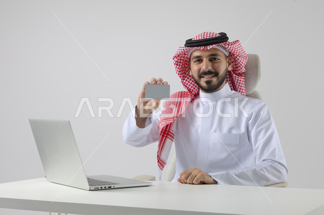عرض موك اب ابيض صغير، مفهوم دمج العمل مع التقنيه والتكنولوجيا, إنجاز مهام العمل عبر جهاز الابتوب, بورتريه لرجل عربي خليجي سعودي مبتسم يرتدي الثوب والشماغ التقليدي يمسك بيده بطاقة تعريفية، العمل على الحاسب المحمول  بإيماءات السرور, خلفيه بيضاء