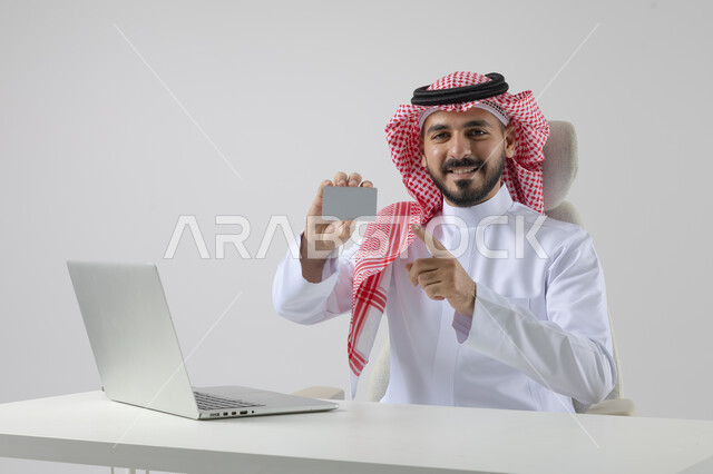 عرض موك اب ابيض صغير، مفهوم دمج العمل مع التقنيه والتكنولوجيا, إنجاز مهام العمل عبر جهاز الابتوب, بورتريه لرجل عربي خليجي سعودي مبتسم يرتدي الثوب والشماغ التقليدي يمسك بيده بطاقة تعريفية، العمل على الحاسب المحمول  بإيماءات السرور, خلفيه بيضاء