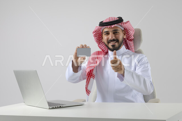 Small white mockup, concept of integrating work with technology, completing work tasks via laptop, portrait of a smiling Saudi Arabian Gulf man wearing traditional thobe and shemagh holding an ID card, working on laptop with gestures of pleasure, white background