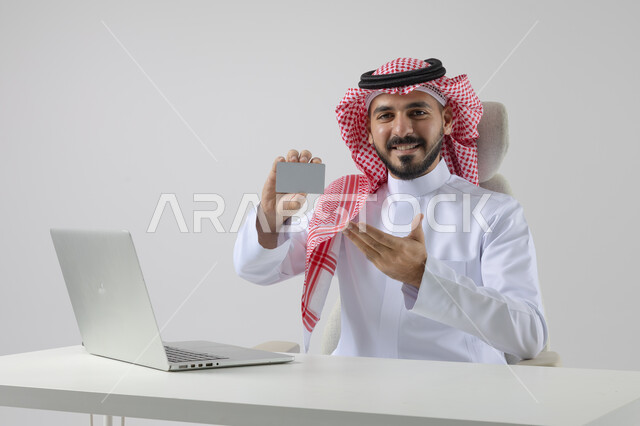 Small white mockup, concept of integrating work with technology, completing work tasks via laptop, portrait of a smiling Saudi Arabian Gulf man wearing traditional thobe and shemagh holding an ID card, working on laptop with gestures of pleasure, white background