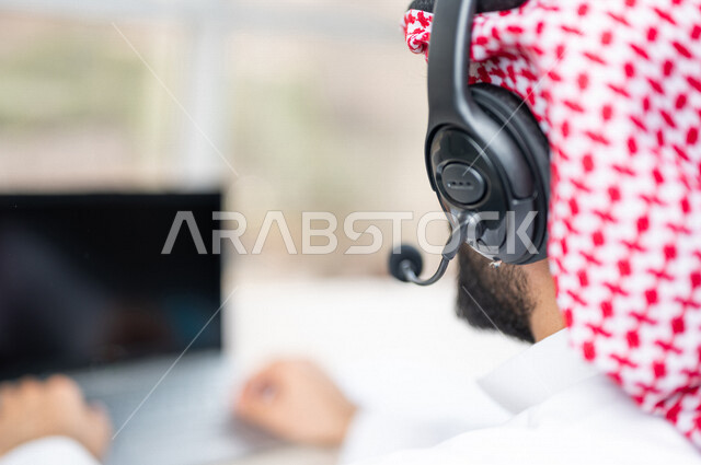 صورة مقربة ليد رجل عربي خليجي سعودي يعمل في خدمة العملاء، موظف يرتدي سماعة الاتصال، استعلامات واستفسارات، الاجابه على اسئلة العملاء، عقد اجتماع عمل عبر الانترنت، العمل عن بعد من خلال الحاسوب المحمول، متابعة وتطوير العمل