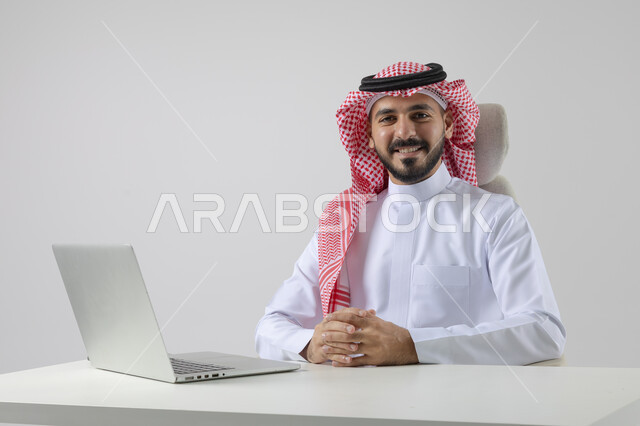 The concept of integrating work with technology, completing work tasks via laptop, portrait of a smiling Saudi Arabian Gulf man wearing the traditional thobe and shemagh working on the laptop with gestures of pleasure, white background