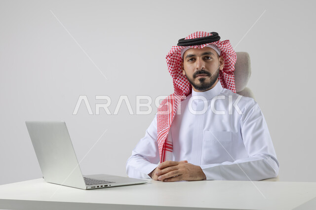 The concept of integrating work with technology, completing work tasks via laptop, portrait of a smiling Saudi Arabian Gulf man wearing the traditional thobe and shemagh working on the laptop with gestures of pleasure, white background