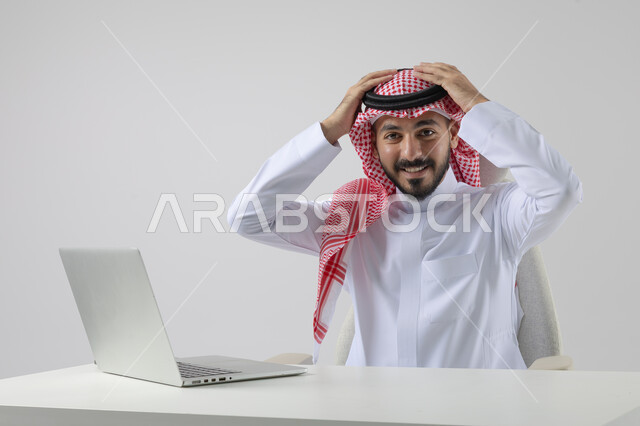 The concept of integrating work with technology, completing work tasks via laptop, portrait of a smiling Saudi Arabian Gulf man wearing the traditional thobe and shemagh working on the laptop with gestures of pleasure, white background