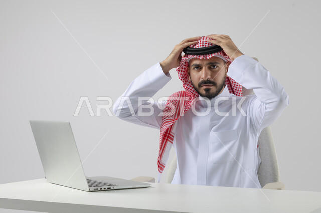  مفهوم دمج العمل مع التقنيه والتكنولوجيا, إنجاز مهام العمل عبر جهاز الابتوب, بورتريه لرجل عربي خليجي سعودي مبتسم يرتدي الثوب والشماغ التقليدي يعمل على الحاسب المحمول  بإيماءات السرور, خلفيه بيضاء