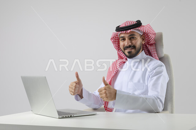  تعابير الاعجاب والقبول، مفهوم دمج العمل مع التقنيه والتكنولوجيا, إنجاز مهام العمل عبر جهاز الابتوب, بورتريه لرجل عربي خليجي سعودي مبتسم يرتدي الثوب والشماغ التقليدي يعمل على الحاسب المحمو بإيماءات السرور, خلفيه بيضاء 