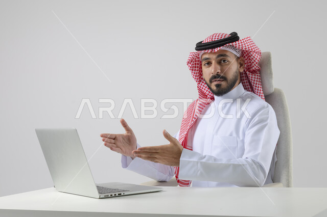 Pointing to laptop, concept of integrating work with technology, accomplishing work tasks via laptop, portrait of a smiling Saudi Arabian Gulf man wearing traditional thobe and shemagh working on laptop with gestures of pleasure, white background