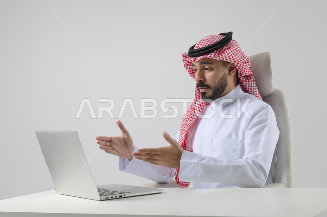 Pointing to laptop, concept of integrating work with technology, accomplishing work tasks via laptop, portrait of a smiling Saudi Arabian Gulf man wearing traditional thobe and shemagh working on laptop with gestures of pleasure, white background