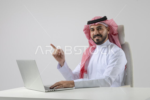 Pointing to laptop, concept of integrating work with technology, accomplishing work tasks via laptop, portrait of a smiling Saudi Arabian Gulf man wearing traditional thobe and shemagh working on laptop with gestures of pleasure, white background