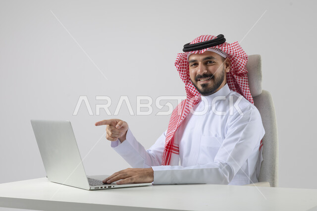 Pointing to laptop, concept of integrating work with technology, accomplishing work tasks via laptop, portrait of a smiling Saudi Arabian Gulf man wearing traditional thobe and shemagh working on laptop with gestures of pleasure, white background