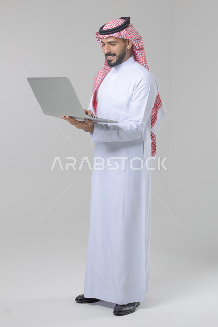 استخدام التقنية الحديثه المتطورة في الحياة العملية، تعابير وايماءات الاندماج والسرور، بورتريه لشاب عربي خليجي سعودي مبتسم يرتدي الثوب التقليدي والشماغ يقوم بالعمل على الحاسب المحمول، صورة الجسم بالطول الكامل، خلفية بيضاء