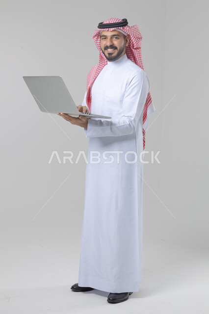 استخدام التقنية الحديثه المتطورة في الحياة العملية، تعابير وايماءات الاندماج والسرور، بورتريه لشاب عربي خليجي سعودي مبتسم يرتدي الثوب التقليدي والشماغ يقوم بالعمل على الحاسب المحمول، صورة الجسم بالطول الكامل، خلفية بيضاء