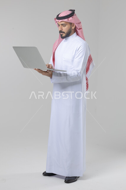 استخدام التقنية الحديثه المتطورة في الحياة العملية، تعابير وايماءات الاندماج والسرور، بورتريه لشاب عربي خليجي سعودي مبتسم يرتدي الثوب التقليدي والشماغ يقوم بالعمل على الحاسب المحمول، صورة الجسم بالطول الكامل، خلفية بيضاء
