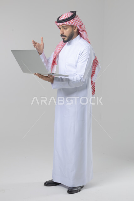 استخدام التقنية الحديثه المتطورة في الحياة العملية، تعابير وايماءات الاندماج والسرور، بورتريه لشاب عربي خليجي سعودي مبتسم يرتدي الثوب التقليدي والشماغ يقوم بالعمل على الحاسب المحمول، صورة الجسم بالطول الكامل، خلفية بيضاء
