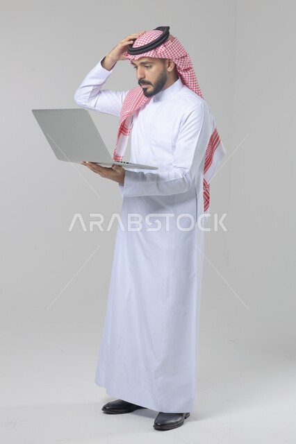 Using modern advanced technology in practical life, expressions and gestures of integration and pleasure, portrait of a smiling young Saudi Arabian Gulf man wearing traditional thobe and shemagh working on a laptop, full body image, white background