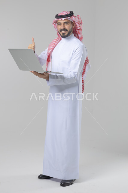 Using modern advanced technology in practical life, expressions and gestures of integration and pleasure, portrait of a smiling young Saudi Arabian Gulf man wearing traditional thobe and shemagh working on a laptop, full body image, white background