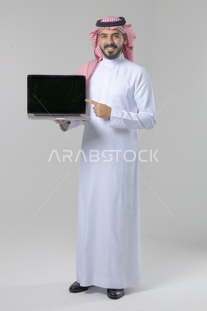 Blank black screen, use of modern advanced technology in practical life, expressions and gestures of admiration and pleasure, portrait of a smiling Saudi Arabian Gulf young man wearing traditional thobe and shemagh working on laptop, full body image, white background