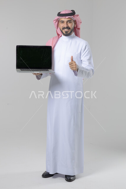 عرض شاشة سوداء فارغة، استخدام التقنية الحديثه المتطورة في الحياة العملية، تعابير وايماءات الاعجاب والسرور، بورتريه لشاب عربي خليجي سعودي مبتسم يرتدي الثوب التقليدي والشماغ يقوم بالعمل على الحاسب المحمول، صورة الجسم بالطول الكامل، خلفية بيضاء