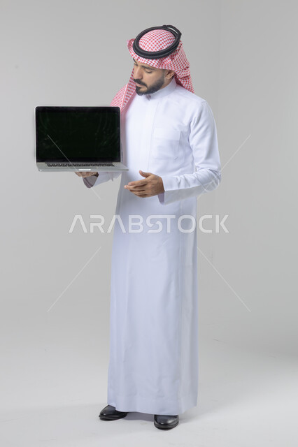 عرض شاشة سوداء فارغة، استخدام التقنية الحديثه المتطورة في الحياة العملية، تعابير وايماءات الاعجاب والسرور، بورتريه لشاب عربي خليجي سعودي مبتسم يرتدي الثوب التقليدي والشماغ يقوم بالعمل على الحاسب المحمول، صورة الجسم بالطول الكامل، خلفية بيضاء