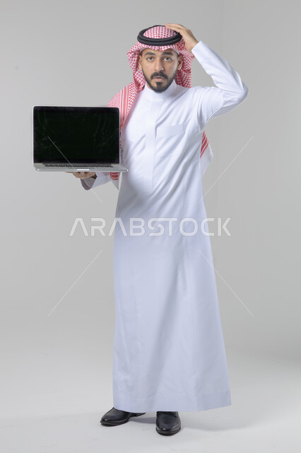 عرض شاشة سوداء فارغة، استخدام التقنية الحديثه المتطورة في الحياة العملية، تعابير وايماءات الاعجاب والسرور، بورتريه لشاب عربي خليجي سعودي مبتسم يرتدي الثوب التقليدي والشماغ يقوم بالعمل على الحاسب المحمول، صورة الجسم بالطول الكامل، خلفية بيضاء