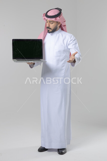 عرض شاشة سوداء فارغة، استخدام التقنية الحديثه المتطورة في الحياة العملية، تعابير وايماءات الاعجاب والسرور، بورتريه لشاب عربي خليجي سعودي مبتسم يرتدي الثوب التقليدي والشماغ يقوم بالعمل على الحاسب المحمول، صورة الجسم بالطول الكامل، خلفية بيضاء