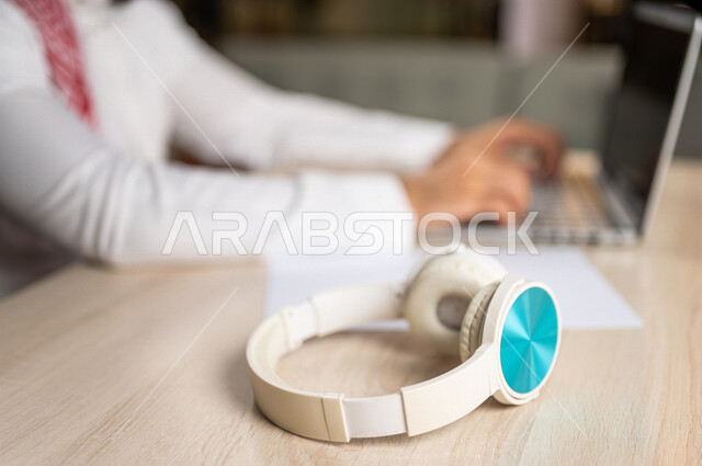 صورة مقربة ليد رجل عربي خليجي سعودي يستخدم الحاسوب المحمول وبجانبه سماعة لاسلكية، الاستماع الى الدورات الالكترونية، الاستماع الى الموسيقى، استخدام تقنيات حديثة