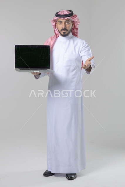 عرض شاشة سوداء فارغة، استخدام التقنية الحديثه المتطورة في الحياة العملية، تعابير وايماءات الاعجاب والسرور، بورتريه لشاب عربي خليجي سعودي مبتسم يرتدي الثوب التقليدي والشماغ يقوم بالعمل على الحاسب المحمول، صورة الجسم بالطول الكامل، خلفية بيضاء