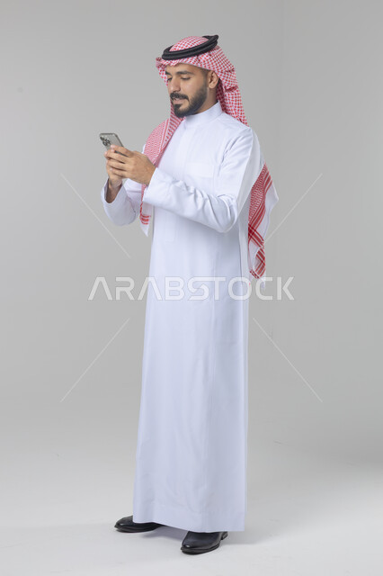  تعابير وايماءات الاندماج والسرور، استخدام التقنية الحديثه المتطورة في الحياة العملية، بورتريه لشاب عربي خليجي سعودي مبتسم يرتدي الشماغ و الثوب التقليدي يقوم بالعمل على الهاتف المحمول، صورة الجسم بالطول الكامل، خلفية بيضاء