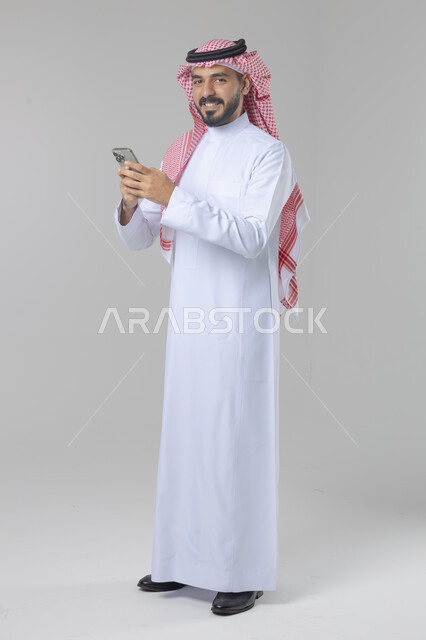  تعابير وايماءات الاندماج والسرور، استخدام التقنية الحديثه المتطورة في الحياة العملية، بورتريه لشاب عربي خليجي سعودي مبتسم يرتدي الشماغ و الثوب التقليدي يقوم بالعمل على الهاتف المحمول، صورة الجسم بالطول الكامل، خلفية بيضاء