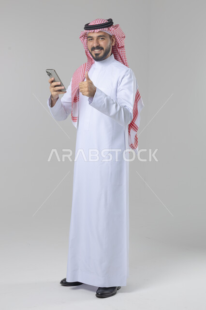  تعابير وايماءات الاندماج والسرور، استخدام التقنية الحديثه المتطورة في الحياة العملية، بورتريه لشاب عربي خليجي سعودي مبتسم يرتدي الشماغ و الثوب التقليدي يقوم بالعمل على الهاتف المحمول، صورة الجسم بالطول الكامل، خلفية بيضاء