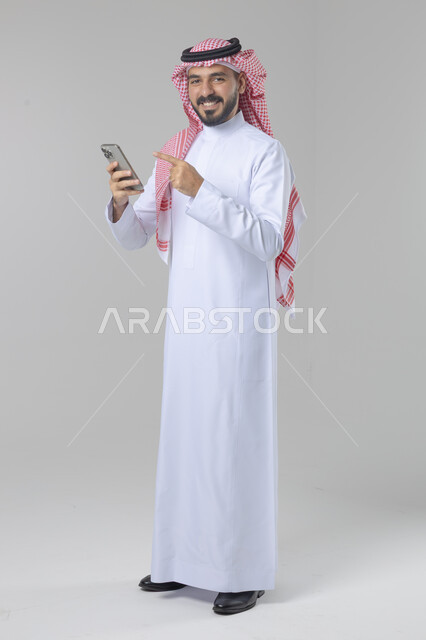  تعابير وايماءات الاندماج والسرور، استخدام التقنية الحديثه المتطورة في الحياة العملية، بورتريه لشاب عربي خليجي سعودي مبتسم يرتدي الشماغ و الثوب التقليدي يقوم بالعمل على الهاتف المحمول، صورة الجسم بالطول الكامل، خلفية بيضاء