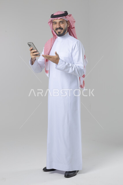  تعابير وايماءات الاندماج والسرور، استخدام التقنية الحديثه المتطورة في الحياة العملية، بورتريه لشاب عربي خليجي سعودي مبتسم يرتدي الشماغ و الثوب التقليدي يقوم بالعمل على الهاتف المحمول، صورة الجسم بالطول الكامل، خلفية بيضاء