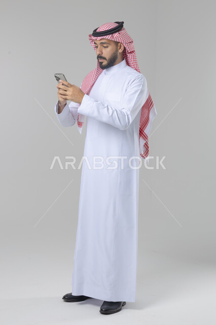  تعابير وايماءات الاندماج والسرور، استخدام التقنية الحديثه المتطورة في الحياة العملية، بورتريه لشاب عربي خليجي سعودي مبتسم يرتدي الشماغ و الثوب التقليدي يقوم بالعمل على الهاتف المحمول، صورة الجسم بالطول الكامل، خلفية بيضاء
